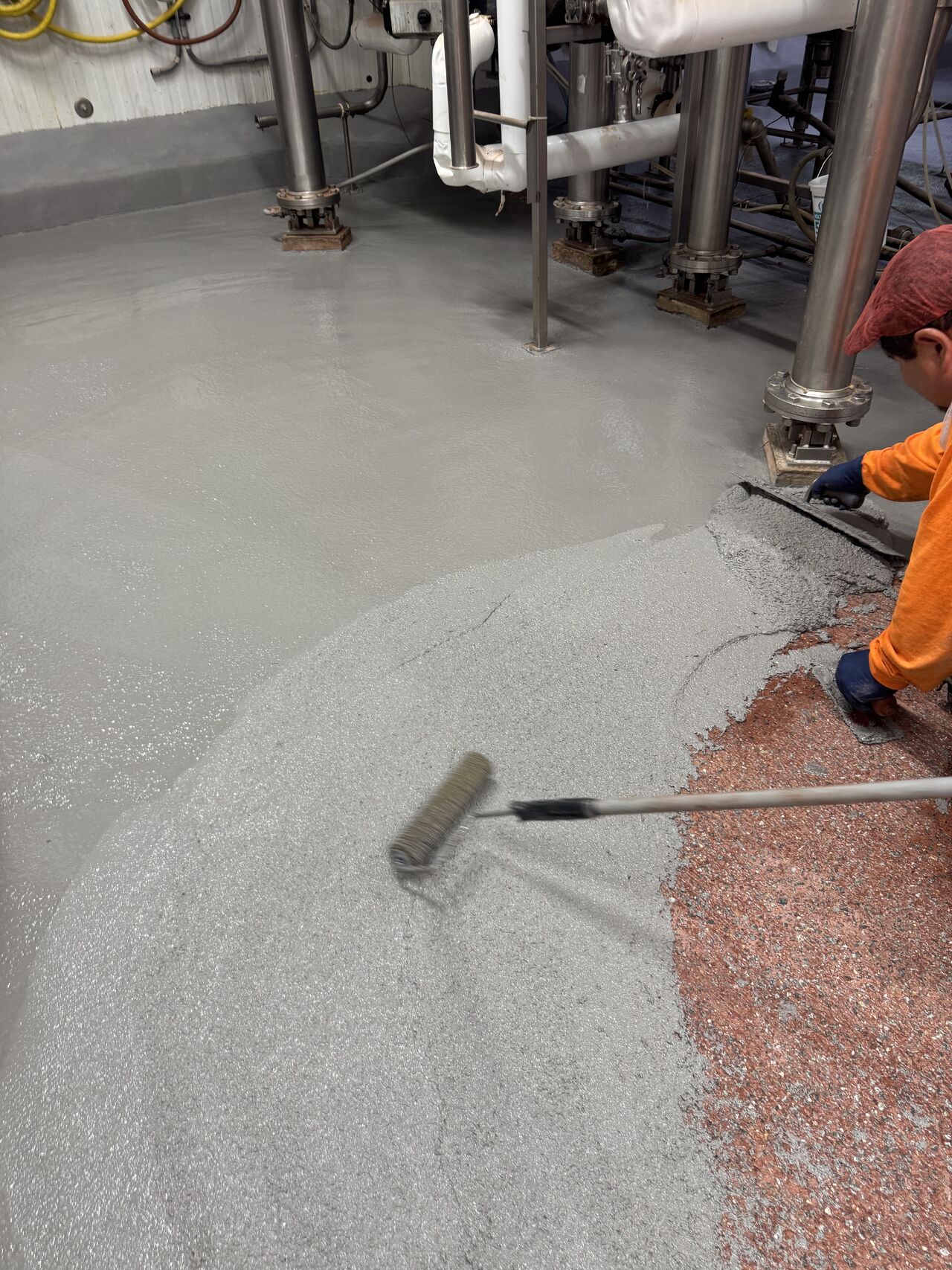 Crew member rolling fresh SaniCrete STX cementitious urethane around pipe pedestals, old red floor visible at edge